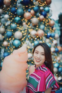 Portrait of smiling young woman standing by christmas tree holding cotton candy outdoors