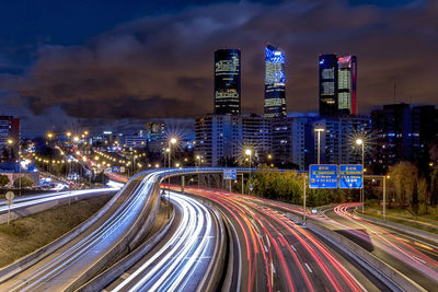 Light trails on road by illuminated buildings against sky at night