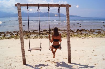 Rear view of woman sitting on swing at beach against sky