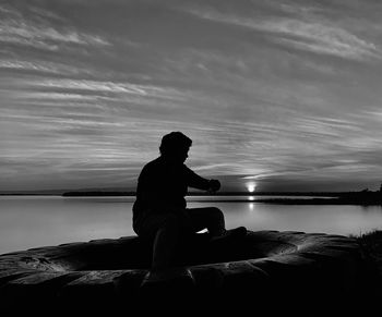 Silhouette man sitting on rock by sea against sky during sunset