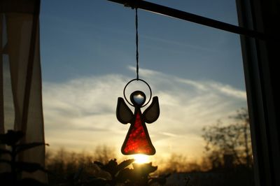 Low angle view of angel figurine hanging on window against sky during sunset