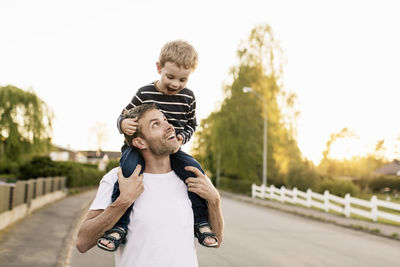 Happy father carrying son on shoulders while standing at street against clear sky