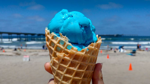 Close-up of hand holding ice cream cone at beach against sky