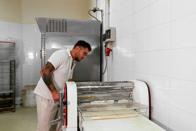 Side view of concentrated male chef in casual clothes and eyeglasses standing and working at dough rolling machine while preparing pastry in professional kitchen