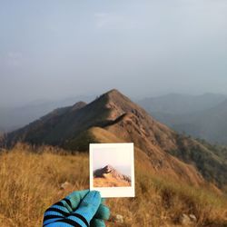 Cropped hand of person holding photograph against mountain and sky