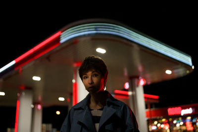 Portrait of style hipster young girl in neon night light standing on street near modern gas station.