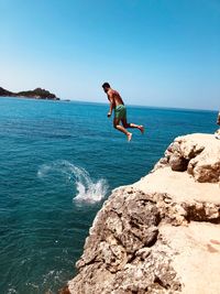 Man jumping on rock in sea against sky