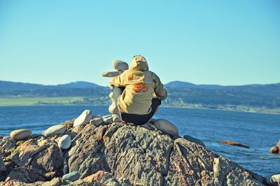 Rear view of man sitting on rock by sea against clear sky