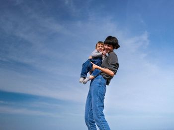 Smiling mother carrying son while standing against blue sky