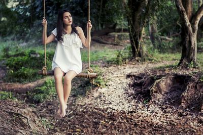 Full length of thoughtful young woman swinging in forest