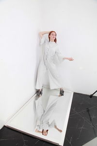 Full length fashion catalog style portrait of woman in jumpsuit standing on mirror over white wall