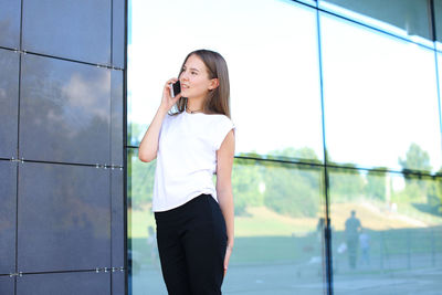 Young businesswoman talking on phone while standing against wall