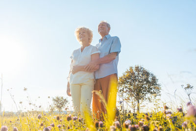 Low angle view of smiling senior couple looking away while standing on field against sky during sunny day