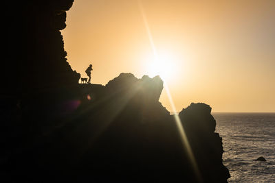 Silhouette person on rock by sea against sky during sunset