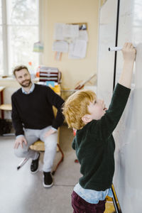 Blond boy writing on whiteboard near male teacher sitting in classroom