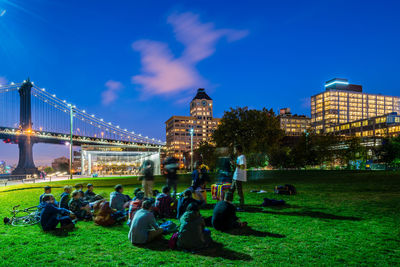 People sitting in park by bridge and illuminated buildings against blue sky at dusk