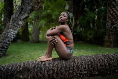 Full body side view of african american female in swimwear looking up while sitting on tree trunk in tropical country