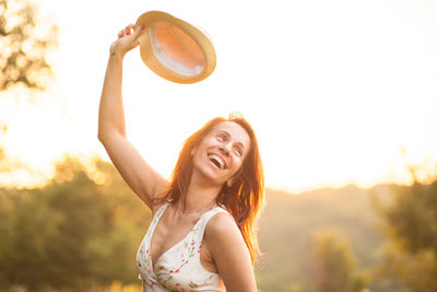 Portrait of a smiling young woman with arms raised against sky during sunset