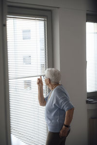 Senior woman looking outside through window blinds at home
