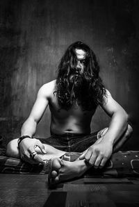 Full length of shirtless man with long hair sitting against wall