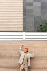 Overwhelmed male in casual outfit yelling and lifting fists in excitement while standing with eyes closed against masonry wall