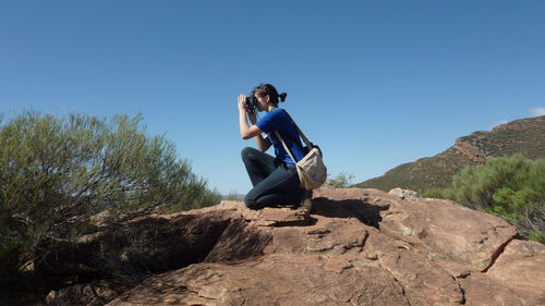 Woman photographing through camera while kneeling on rock against clear sky