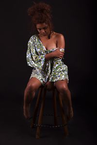 Full length of sensuous young woman sitting on stool against black background