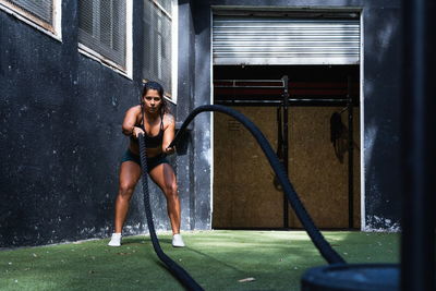 Full body of determined hispanic sportswoman in black activewear doing exercise with battle ropes during intense workout in light gym