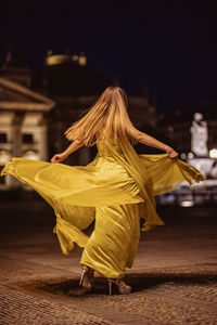 Woman wearing yellow evening gown while spinning on footpath in city at night