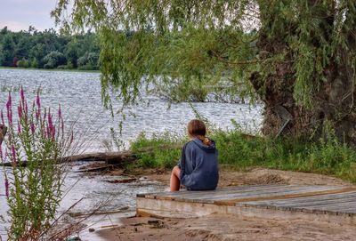 Rear view of young lady sitting by lake against trees