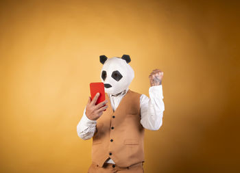 Man in panda bear mask on yellow background using smartphone.