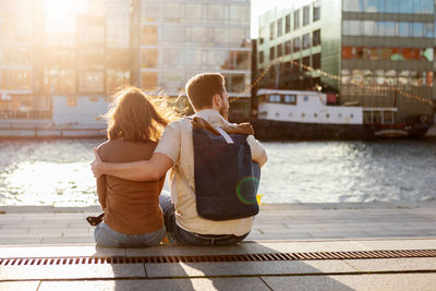 Rear view of man sitting with arm around woman against river in city