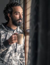 Bearded man holding coffee cup while looking through window