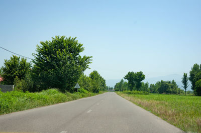 Empty road along trees on field against clear sky
