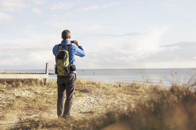 Rear view of hiker photographing sea through smart phone at beach