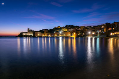 Illuminated buildings by sea against sky at night