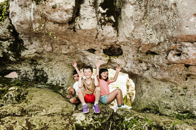 Full body happy boys and girls in casual clothes raising arms and smiling while sitting near rough rock formation on sunny summer day