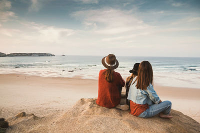 Female friends sitting at beach against sky during sunset