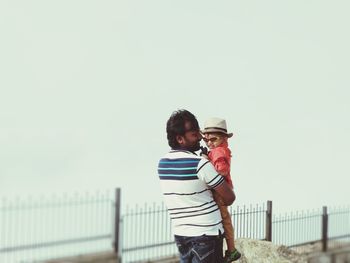 Smiling man with son standing by fence against clear sky