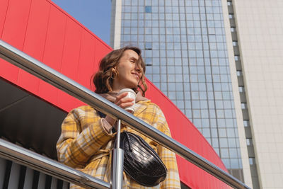 Happy caucasian woman in a yellow coat with cup of coffee to go on city walk.