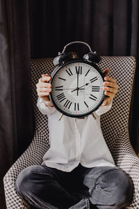 Businesswoman covering her face with vintage analog clock sitting in the black and white chair. 