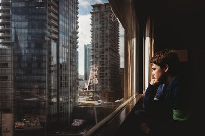 Tween boy looking out a window at tall buildings of the city outside.