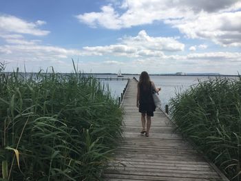 Full length rear view of woman walking on pier amidst plants against sea and sky