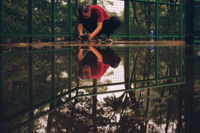 Man photographing puddle through mobile phone while crouching by fence