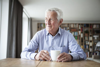 Portrait of pensive senior man sitting at table with cup of coffee looking through window
