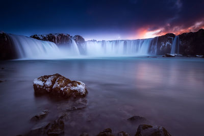 Scenic view of waterfall against cloudy sky at sunset