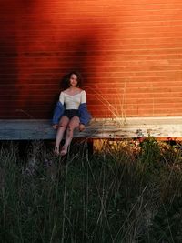 Portrait of smiling young woman sitting on wood against brick wall