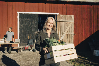 Portrait of smiling mature woman carrying crate full of vegetables with barn in background
