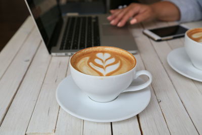 Cropped image of person using laptop by cappuccino served on table