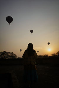 Rear view of woman standing by wall against sky with hot air balloon during sunset